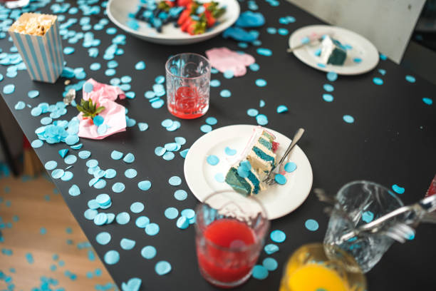 A gender reveal party table scattered with blue confetti, indicating a baby boy. Cake with blue layers, drinks, and snacks are visible, set in an indoor setting. The joyous occasion is marked by festive decorations and casual dining ware.