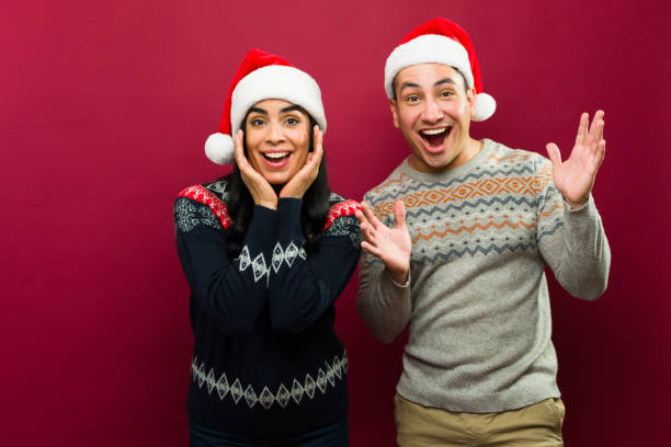 istock Attractive young couple in festive sweaters and santa hats posing in a studio setting and looking excited 2166526901