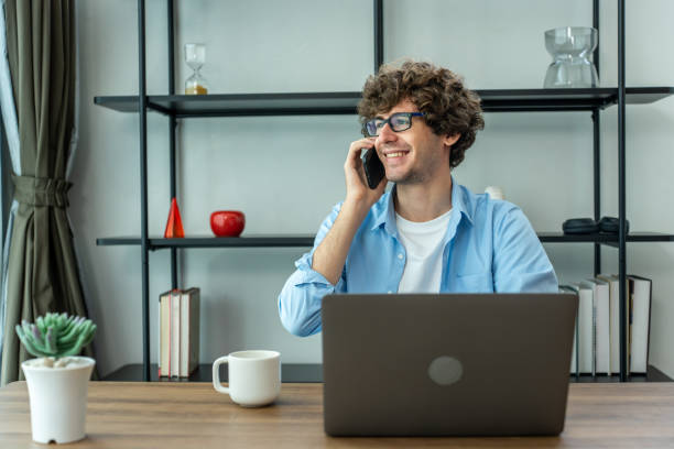 istock Caucasian young businessman talking on phone while working from home. Attractive handsome freelancer guy sitting on table and talking on smartphone to communicate with coworker in living room at house 2166570841