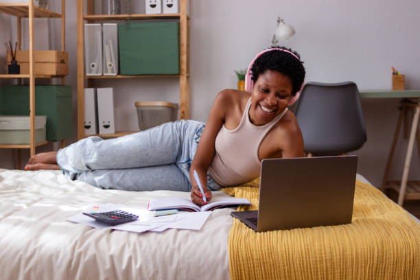 istock Young black woman lying on bed taking notes while watching online class on laptop 2166571938