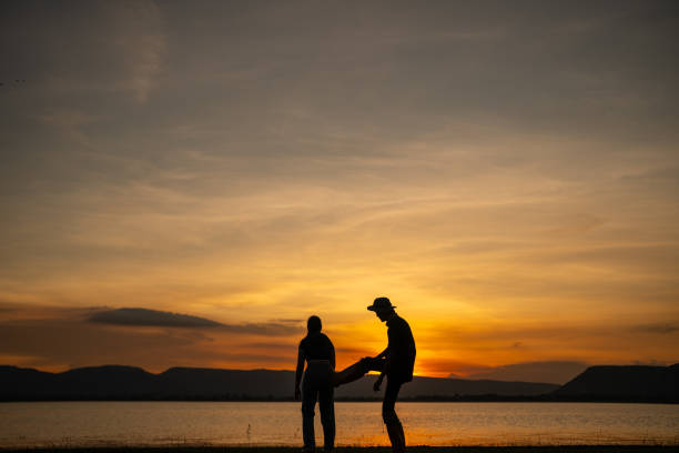 Two silhouettes of people standing on a beach at sunset. The man on the left is holding a bag