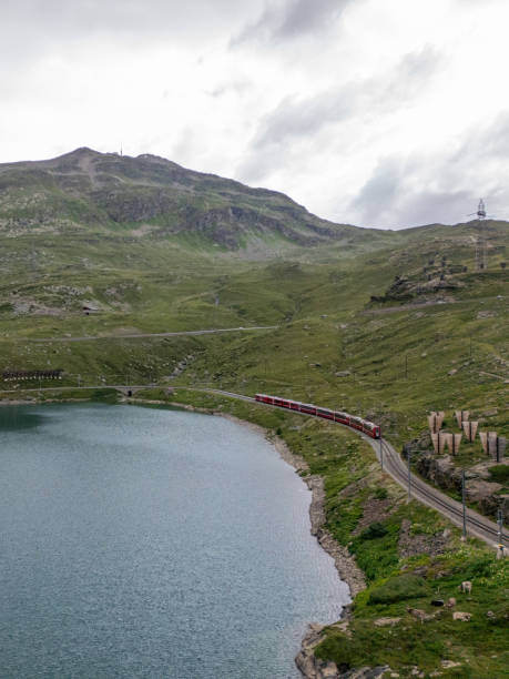 istock Red Train Curving Alongside Mountain Lake on Cloudy Day 2166573648
