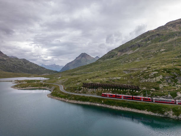 istock Red Train Traveling Along Mountain Lake Under Cloudy Skies 2166574180