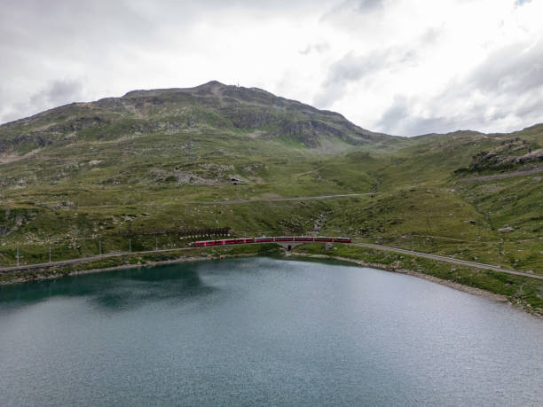 istock Red Train Curving Alongside Mountain Lake on Cloudy Day 2166574194