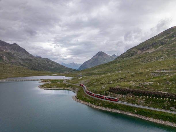 istock Red Train Traveling Along Mountain Lake Under Cloudy Skies 2166574230