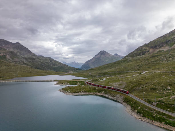 istock Red Train Traveling Along Mountain Lake Under Cloudy Skies 2166574256