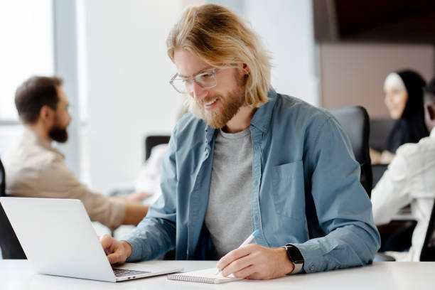 istock Smiling young man taking notes while working on laptop in busy office 2166579527