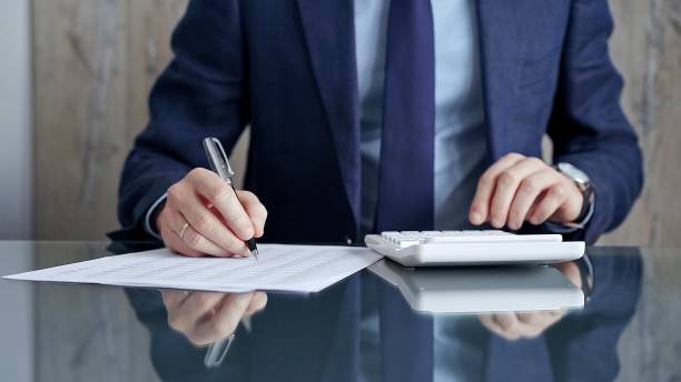 istock Businessman or auditor analyzing financial documents with magnifying glass and calculator at his glass office desk in formal blue jacket. Business people concept 2166588366