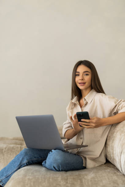 istock Smiling young woman using mobile phone while sitting on a couch at home with laptop computer 2166589445