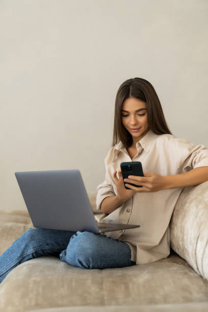 istock Smiling young woman using mobile phone while sitting on a couch at home with laptop computer 2166590344