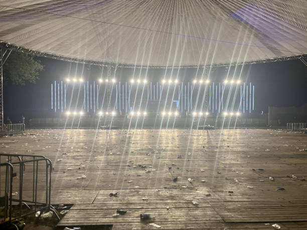 This image shows an empty festival site after an event. The large, open space under a stretched tent canopy is illuminated by bright stage lights in the background. The wooden floor is littered with trash, such as plastic cups and bottles, indicating a recently concluded music festival or concert. The atmosphere is calm, almost deserted, after an evening of hustle and celebration.