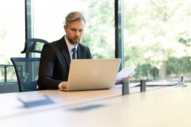 istock Mature businessman with beard working with documents, contracts and bills sitting at table using laptop at work in modern bright office with large windows 2166595156