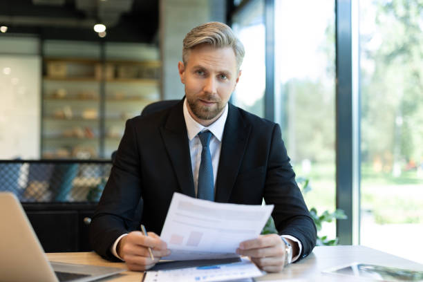 istock Mature businessman with beard working with documents, contracts and bills sitting at table using laptop at work in modern bright office with large windows 2166595199