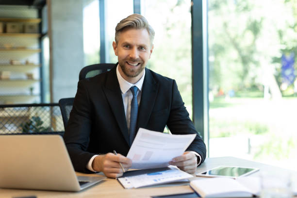 istock Mature businessman with beard working with documents, contracts and bills sitting at table using laptop at work in modern bright office with large windows 2166595207