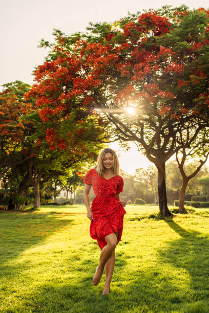 A woman in a vibrant red dress radiates happiness as she strolls through a sun-drenched garden. The lush green lawn beneath her feet contrasts beautifully with the bright blossoms of the trees above, creating a vivid and joyful scene. The sunlight filters through the leaves, casting a warm, golden glow that enhances the carefree atmosphere. Her light-hearted posture and the flow of her dress in the gentle breeze evoke a sense of freedom and connection with nature. This image captures the essence of summer's joy and the simple pleasure of being immersed in nature's beauty.