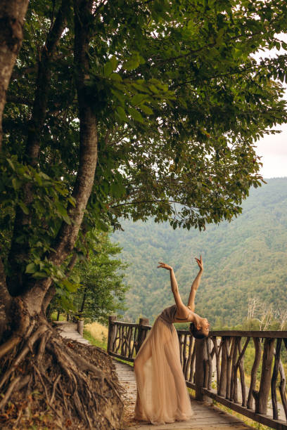 A serene photograph capturing a graceful woman dancing on a wooden path surrounded by lush greenery. Her elegant pose and flowing dress blend harmoniously with the natural landscape, creating a tranquil and enchanting scene. The backdrop of rolling hills and trees adds to the peaceful ambiance, highlighting the beauty of movement and nature. This image epitomizes the essence of freedom, serenity, and the connection between human and environment.