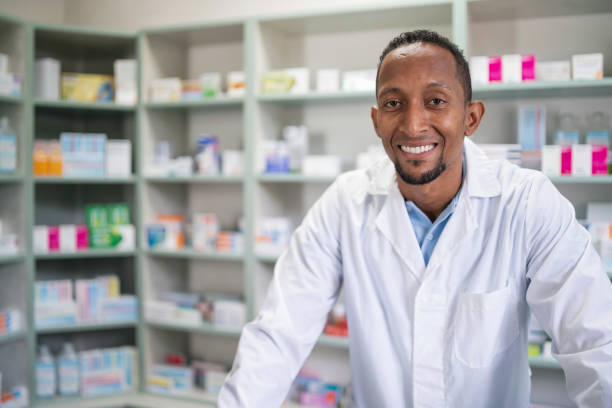 istock Professional confident black pharmacist wearing lab coat, looking at the camera and smiling.  Druggist in drugstore with shelves with health care products 2166601582