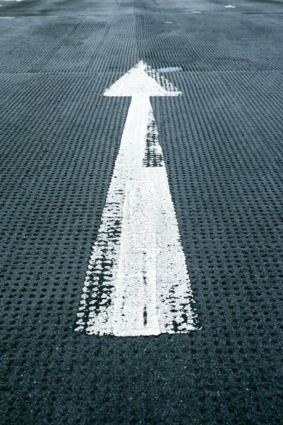 High-resolution image of a straight directional arrow painted on an asphalt road, symbolizing forward direction and navigation. Ideal for projects related to road safety, travel, transportation, and guidance. The bold white arrow contrasts sharply against the dark road surface, capturing the essence of clear, decisive direction.