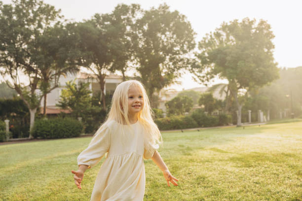 A happy 5-year-old European girl with blonde hair in a red sundress and a white T-shirt jumps on the spot on the grass