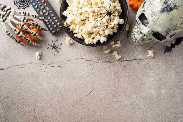 istock Halloween-themed setup featuring a bowl of popcorn, remote control, skeleton hand, skull, and spider decorations on a concrete background 2166615927