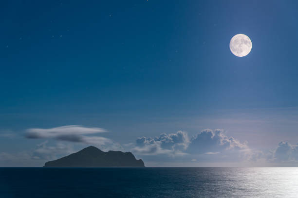 The full moon illuminates the tranquil night view of Turtle Island. The sky was filled with dramatic clouds in varying shades of purple and blue. Touzhen, Yilan County, Taiwan.