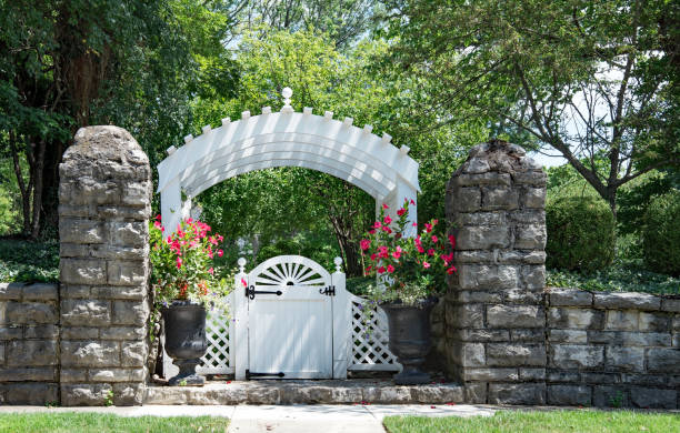 Old, flagstone, garden wall with white, arched gate and red Mandevilla flowers in urns in a residential neighborhood.