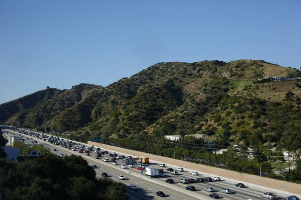 The volume of traffic on the road in Los Angeles, California during the summer. In the afternoon under a blue sky, workers drive on the highway after work. The background shows green mountains.