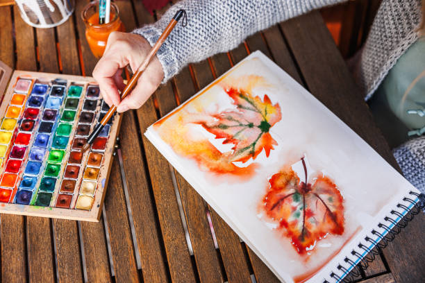 Woman artist dips paintbrush into color palette and painting a watercolor illustration