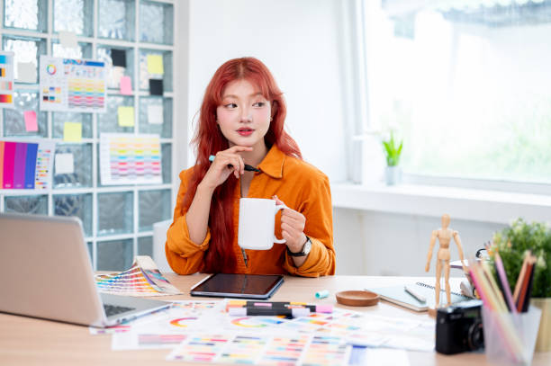 istock A creative Asian female graphic designer is holding a coffee mug, looking away thoughtfully. 2166701561