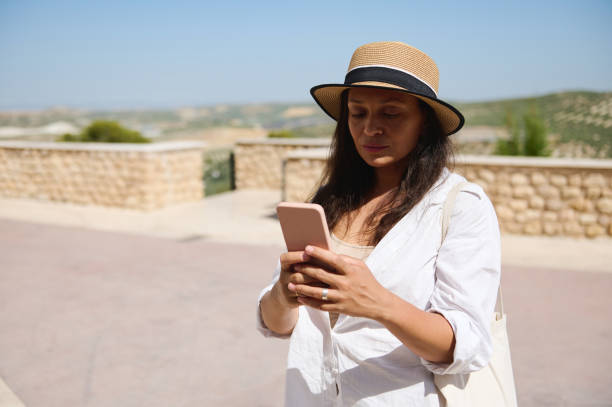 istock Woman using smartphone outdoors on a sunny day, dressed in a hat and white shirt 2166704330