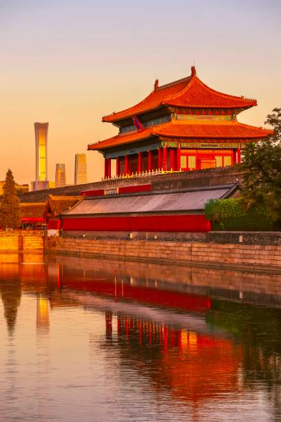 The Gate of Divine Prowess (Shenwu Gate) at the Forbidden City in Beijing is bathed in the warm hues of sunset, with its reflection beautifully cast on the surrounding water. In the background, modern skyscrapers including the CITIC Tower rise, creating a striking visual contrast between Beijings historical and contemporary architecture.