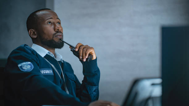African American security officer works in monitoring center during night shift African American security officer or dispatch in uniform uses tablet computer and walkie talkie, looks at screens working in monitoring center during night shift. Surveillance and CCTV system concept. professional guard services stock pictures, royalty-free photos & images