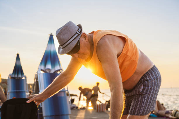 A senior family member carries folded beach chairs away from the sandy shore at sunset, with other family members visible in the background, signaling the peaceful close of a summer day by the sea.