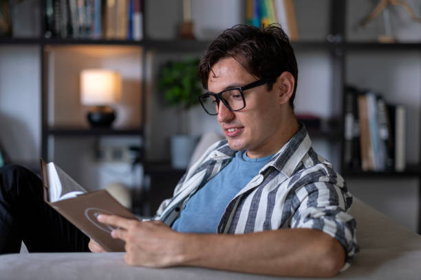 istock Young man with glasses reading a book at home 2166723400