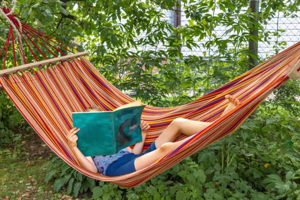 An unrecognizable boy lying in hammock reading book in garden on sunny summer afternoon.