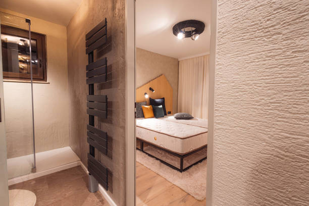 A modern bedroom with a cozy atmosphere, featuring a bed with decorative pillows, a stylish headboard, and a warm color palette. The room is partially visible through a mirror, showcasing a shower area with glass doors.