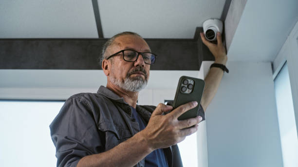 istock Male mature technician sets up CCTV camera in office building using mobile phone 2166740611
