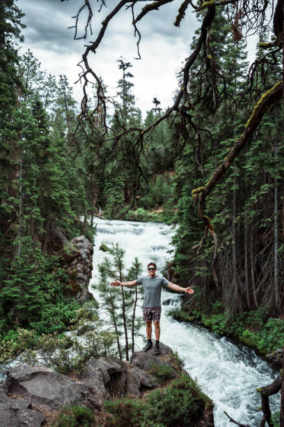 A fit male hiker poses with his arms stretched wide, standing on a rock in front of beautiful river rapids in a Central Oregon park.
