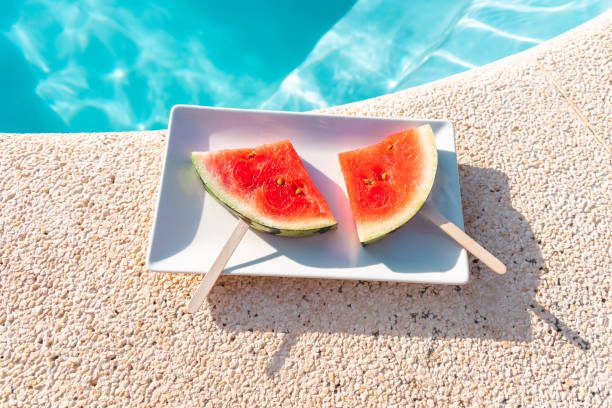 Refreshing watermelon slices on sticks placed on a plate by the edge of a bright blue swimming pool.