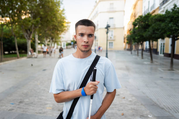 Young blind man is holding a white cane and wearing a blue wristband while walking in the city