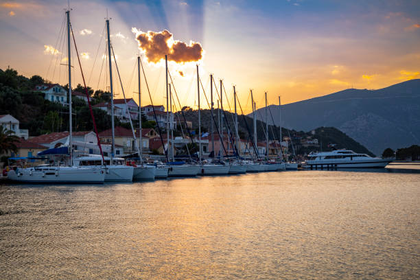A stunning sunset illuminates the harbour in Vathi, Greece, casting a golden glow over the calm waters. The picturesque village is nestled between rolling hills, with boats gently anchored in the bay. Dramatic clouds add depth to the sky, creating a tranquil yet captivating scene as day transitions into night on the island of Ithaca.
