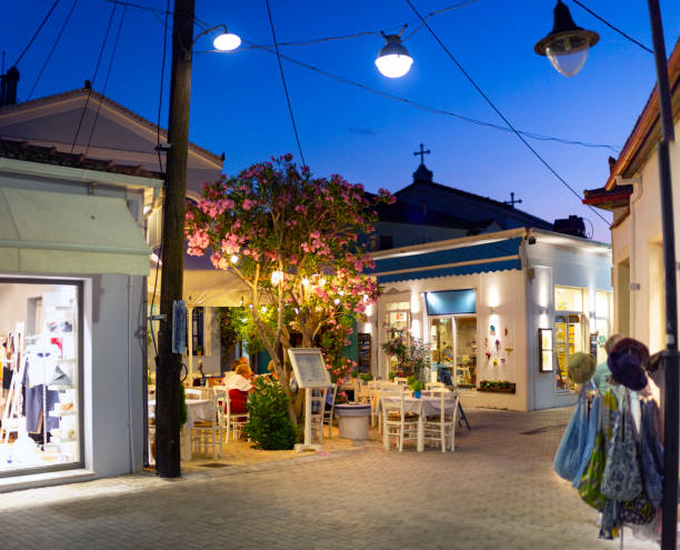 A cosy evening scene in Vathi, Greece, featuring a charming street lined with illuminated shops and outdoor cafes. The soft glow of the streetlights enhances the warm ambience as locals and visitors enjoy the peaceful atmosphere. The deep blue sky transitions into night, highlighting the tranquil allure of this Mediterranean village.