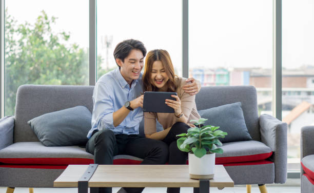 istock Young asian couple sitting on a couch, laughing together and sharing a tablet computer, in a bright living room. 2166824028