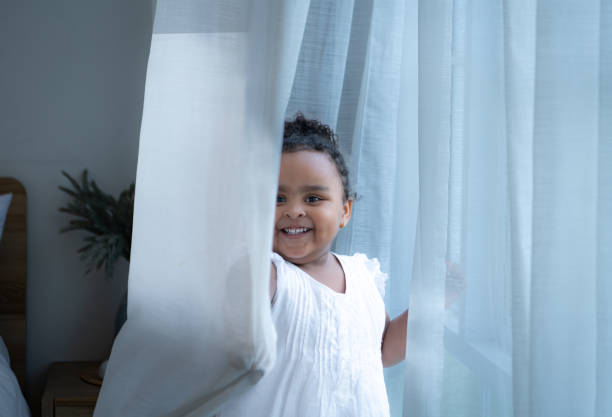 A tiny girl plays hide-and-seek with drapes in her bedroom
