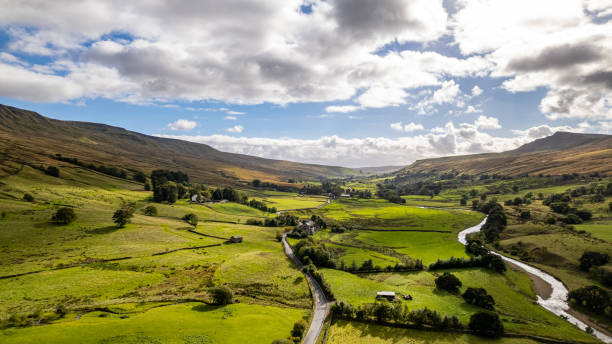 istock Aerial view of a lush green valley with a winding river, patchwork fields, and rolling hills under a partly cloudy sky. 2166831983