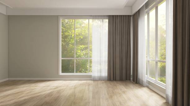 Empty room with gray wall, brown blackout and white sheer curtain in sunlight from backyard with trees on laminated parquet floor
