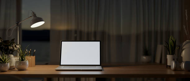 istock A laptop computer on a wooden table by the window in a house at night, illuminated by a table lamp. 2166836541