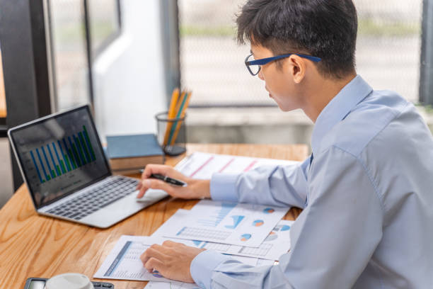 istock Young businessman working on his project in his office checking business result data from the printed sheets and his laptop computer 2166841686