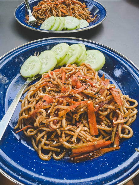 A plate of cooked noodles sits on a floral-patterned, green tablecloth. The green plate features decorative flowers and is filled with noodles mixed with sliced vegetables like bell peppers and onions. The tablecloth's intricate design consists of large floral motifs in gold and cream tones, complementing the vibrant look of the dish. The noodles appear tossed in a light sauce, giving them a slight sheen.