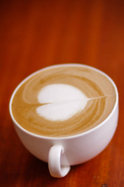 Stock photo showing close-up view of a latte coffee served for breakfast on a cafe table. The coffee has a typically frothy milk foam surface with a heart-shaped pattern latte art design as part of St. Valentine's Day celebrations.